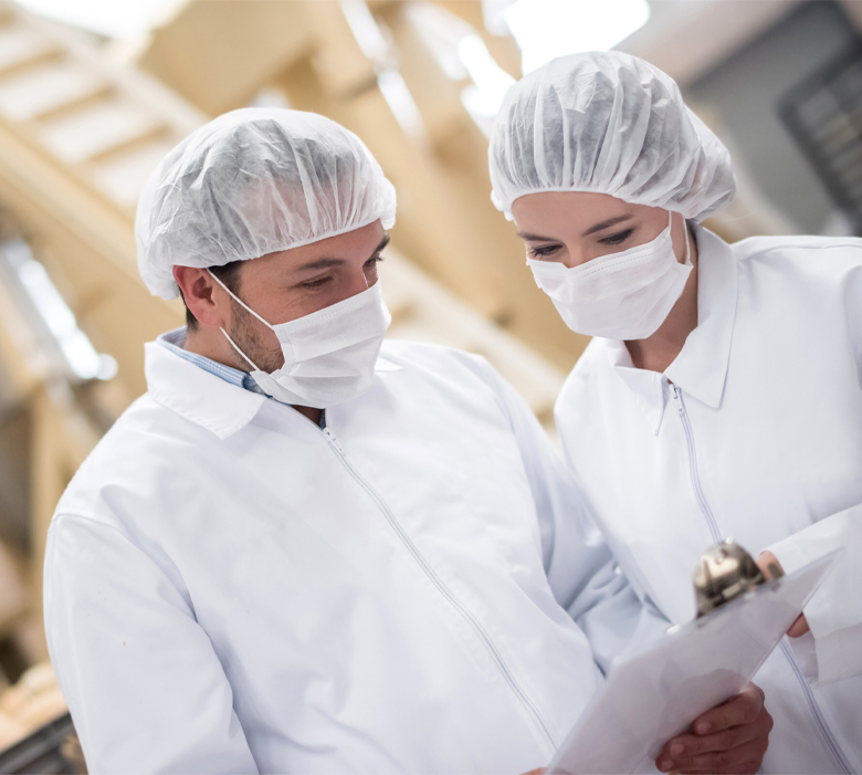 gumbase_Safety QHSE - Gumbase Two factory workers in white protective suits, hair nets, and masks review a clipboard in a clean production area.