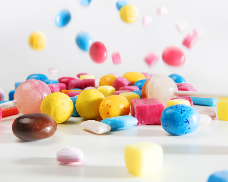 gumbase_Any product format - Gumbase Colorful assortment of candies spilled on a white surface, featuring blue, yellow, pink, and red sweets.