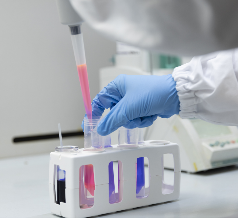Gloved hand uses a pipette to transfer pink liquid into test tubes in a laboratory.