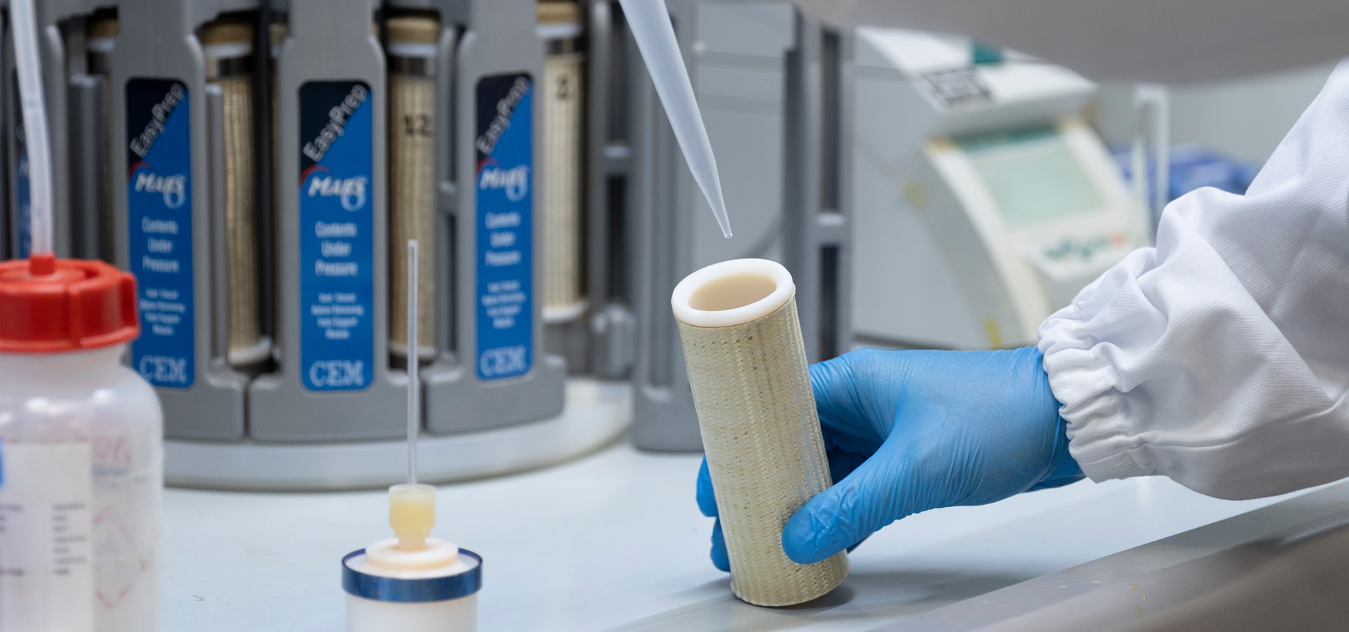 Gloved hand holds a beige cylindrical filter cartridge at a lab bench, a pipette tip hovering above it.