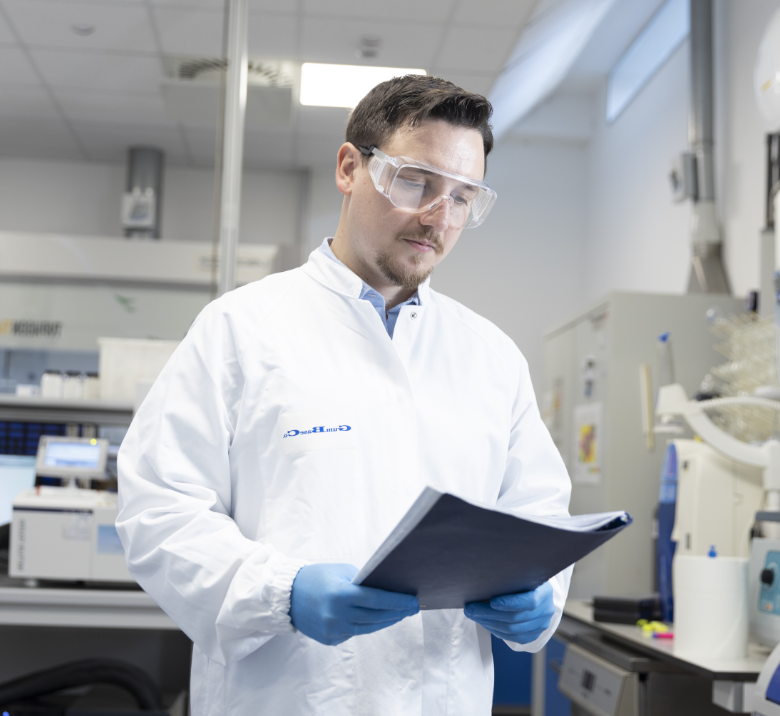 Lab researcher in a white coat and safety goggles reads a clipboard in a modern laboratory
