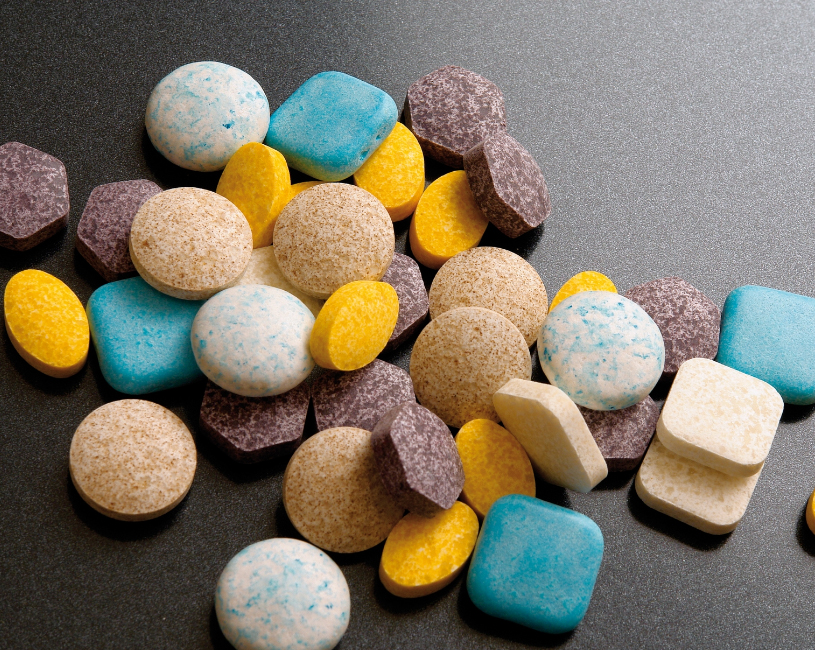 powergum_Compressed tablets - Gumbase Assorted round and square candies or pills in blue, yellow, beige, and purple scattered on a dark surface