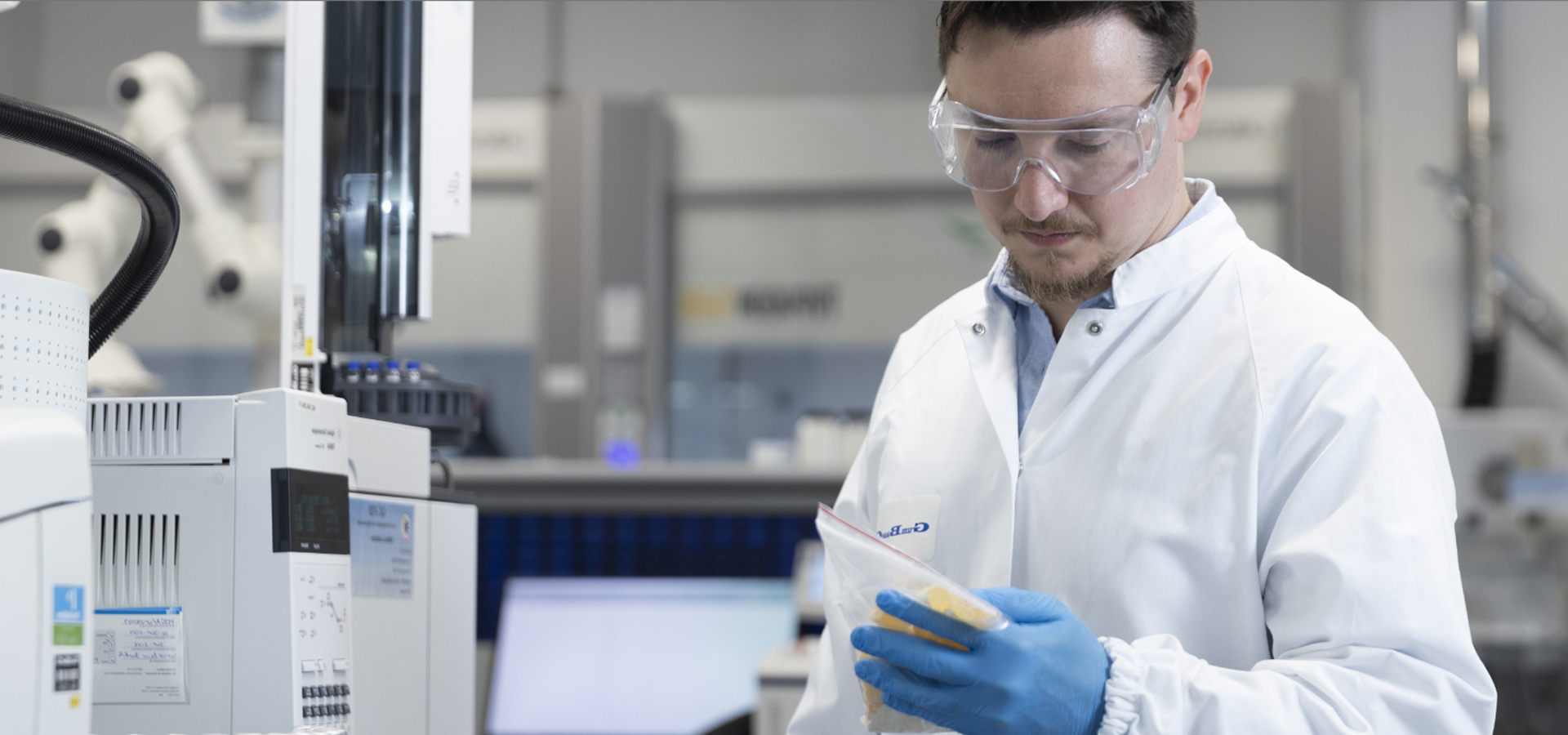 Scientist in a lab coat and safety goggles holds a bag of samples near laboratory equipment.
