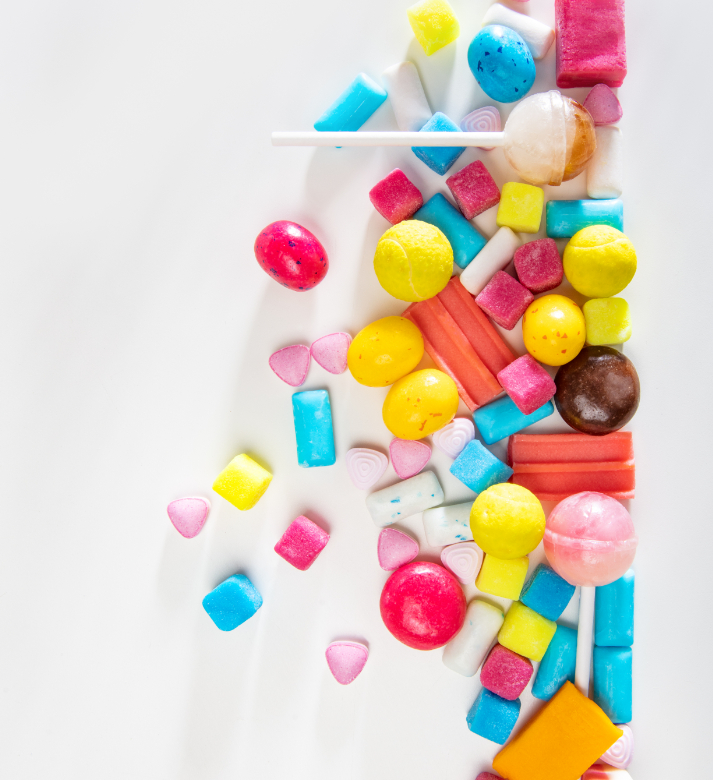 Colorful assortment of candies and lollipops scattered on a white surface, with sugar cubes and gummy shapes scattered throughout, on the right side of the image.